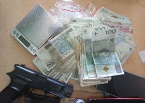 Na blacie stołu leżą pistolet, plik banknotów i waga elektroniczna
