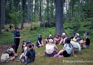 Policjantka mówi do dzieci siedzących na ziemi w kółku, w środku lasu.