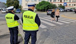 Dwaj policjanci stojący przy przejściu dla pieszych znajdującym się przy szkole. Przez pasy przechodzą dwie dziewczynki.