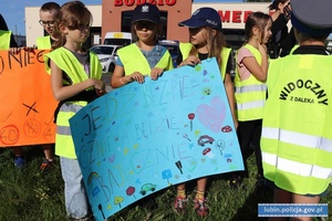 Dzieci w kamizelkach odblaskowych z plakatami z napisami: jedź bezpiecznie a będzie bajecznie.