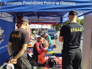 Dwaj strażacy stojący przy namiocie Komendy Powiatowej Policji w Oleśnicy w którym ratownicy medyczni pokazują jak udzielać pierwszej pomocy.