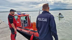 Policjant i ratownik medyczny stojący przy łodzi wodnej służby ratunkowej obserwują zalew i płynącego na skuterze mężczyznę.