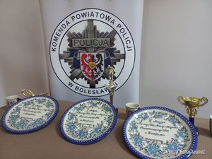 Gwiazda policyjna z napisem Komenda Powiatowa Policji w Bolesławcu i leżące na stole nagrody; puchary i ceramiczne talerze.
