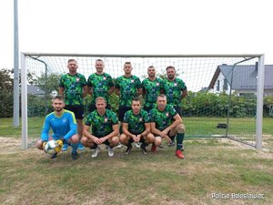 Dziewięciu mężczyzn w strojach sportowych, stojących pod siatką na boisku piłkarskim i pozujący do zdjęcia drużynowego.