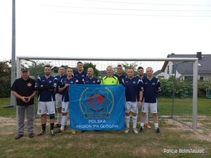 Jedenastu mężczyzn w strojach sportowych, stojących pod siatką na boisku piłkarskim i pozujący do zdjęcia drużynowego, trzymający w ręku plakat z napisem POLSKA Region IPA Głogów.