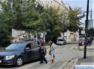 Policjantka stojąca na ulicy podchodzi do samochodu osobowego. Towarzyszy jej kobieta.