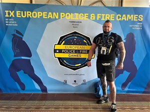 Dobrze zbudowany mężczyzna w stroju sportowym stojący przy banerze z napisami po angielsku 9 European Police &amp; Fire Games
