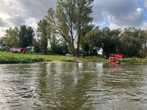Dwa wozy strażackie i łódź strażacka nad brzegiem rzeki.