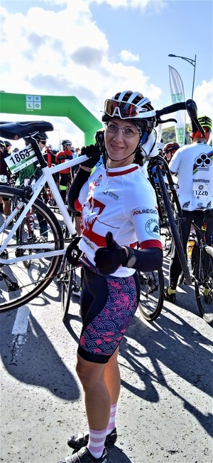 Kobieta w stroju sportowym jako zawodniczka, trzyma na ramieniu rower, stojąc  przed metą.