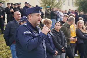 policjant w granatowym mundurze z mikrofonem w ręku, za nim stojące osoby