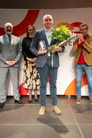 Bartłomiej Majchrzak Pełnomocnik Komendanta Wojewódzkiego Policji we Wrocławiu do spraw Ochrony Praw Człowieka z kwiatami podczas uroczystości z tyłu inni uczestnicy uroczystości bijący mu brawa