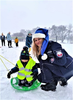 Policjantka kuca na śniegu przy dziecku siedzącym na ślizgu.