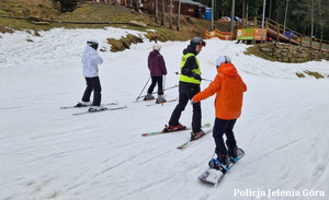 Na stoku. Policjant na nartach pomaga osobie jadącej na snowboardzie. W tle dwie osoby na nartach.