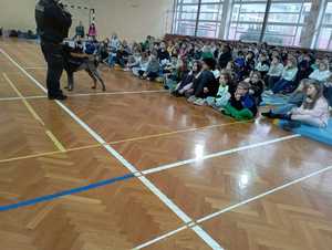 Dzieci na sali gimnastycznej siedzący na podłodze i policjant z psem opowiadający dzieciom o bezpieczeństwie