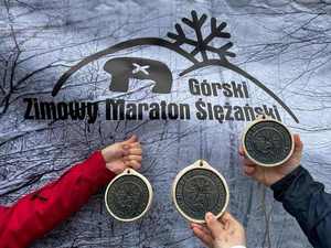 Trzy medale trzymane przez dłonie na tle baneru z napisem Górski Zimowy Maraton Ślężański