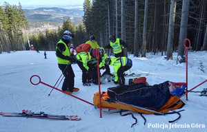 Policjanci w kamizelkach odblaskowych przenoszą poszkodowanego ze śniegu na sanie