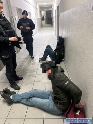 Zatrzymane dwie osoby siedzące na korytarzu na ziemi, nad nimi stoją dwaj policjanci