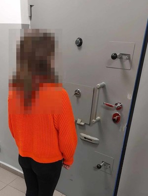 Na zdjęciu widoczna kobieta, która stoi tyłem do osoby wykonującej zdjęcie. Fotografia wykonana w pomieszczeniu zamkniętym na tle drzwi.