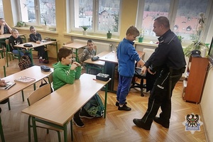 Policjant pokazuje chłopcu technikę samoobrony, reszta klasy siedzi w ławkach i obserwuje