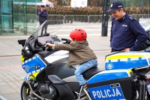 Dziecko siedzące na motocyklu policyjnym, z boku stoi uśmiechnięty policjant