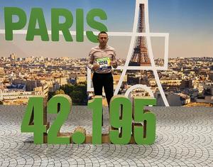 Mężczyzna z numerem startowym pozuje na tle banera z napisem Paris i cyfrą 42.195