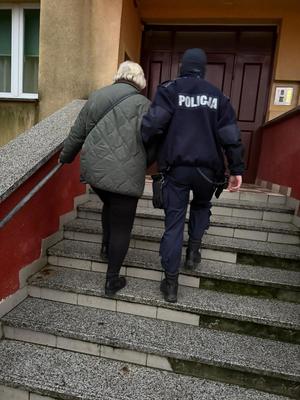 Policjant pomaga starszej kobiecie - prowadzi ją pod rękę po schodach do klatki bloku