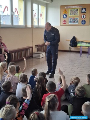 stojący policjant podchodzi z uśmiechem do siedzących na ziemi dzieci