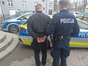 policjant prowadzi do radiowozu zatrzymanego mężczyznę skutego w kajdanki