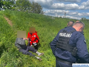ratownik medyczny podaje mężczyźnie siedzącemu na wzgórzu wodę obok policjanci