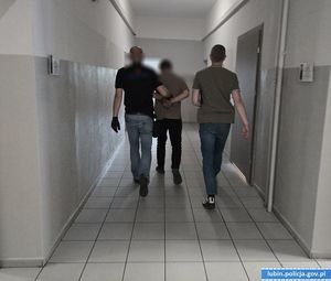 Dwaj policjanci w cywilnych ubraniach prowadzą skutego w kajdanki mężczyznę korytarzami komendy
