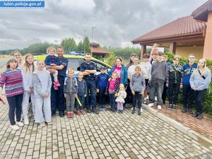 Umundurowani policjanci z dziećmi - uczestnicy półkolonii. Zdjęcie wykonane na tle oznakowanego radiowozu policyjnego