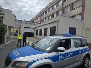 radiowóz przed szpitalem na pierwszym planie, dalej dwaj policjanci stojący tylem