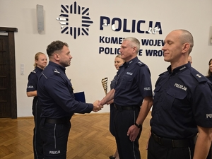 Komendant podaje rękę policjantowi w tle stoją pozostali wyróżnieni policjanci, obok stoi policjant trzymający wcześniej wręczony puchar