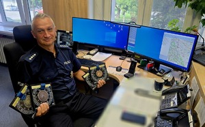 Policjant siedzący przed dwoma monitorami trzymający w rękach medale z zawodów strzeleckich