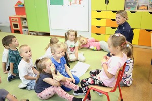 Policjantka siedząca na dywanie razem z dziećmi w budynku przedszkolnym tłumaczy dzieciom zasady bezpieczeństwa.