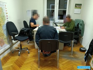 dwaj policjanci siedzący przy biurkach przesłuchują mężczyznę w kajdankach siedzącego na krześle obok