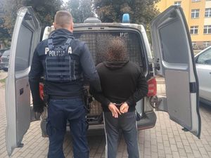 policjant stojący przodem do otwartego radiowozu trzyma mężczyznę zakutego w kajdanki