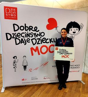 Policjantka stojąca na tle baneru z napisem Dobre Dzieciństwo Daje Dziecku Moc