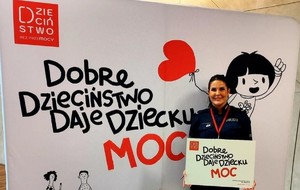 Policjantka stojąca na tle baneru z napisem Dobre Dzieciństwo Daje Dziecku Moc