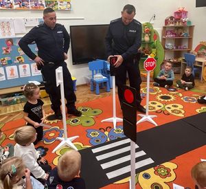 Policjanci pokazują grupie przedszkolaków w sali, jak bezpiecznie przechodzić przez pasy