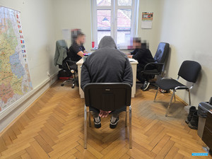 mężczyzna w bluzie z kapturem na głowie siedzi na krześle pomiędzy dwoma przesłuchującymi go policjantami