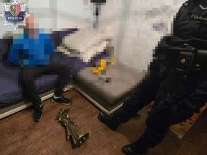 policjant z zatrzymanym mężczyzną siedzącym na łóżku, na ziemi leży figurka „dobrego duszka”