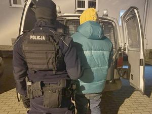 Policjant ubrany w czarny mundur doprowadza do radiowozu mężczyznę ubranego w kurtkę koloru niebieskiego oraz żółtą czapką