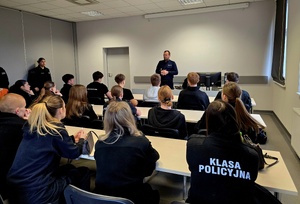 uczniowie klas mundurowych siedzą w ławkach, przemawia do nich policjant