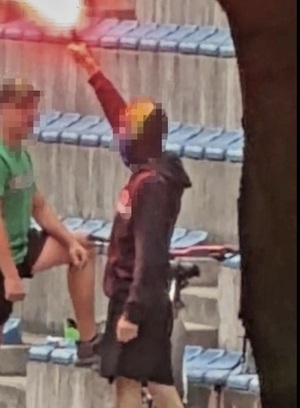 Pseudokibic stoi na trybunie stadionu z uniesioną ręką w której trzyma odpaloną racę świetlną.