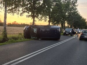 Samochód osobowy znajduje się na boku na drodze. W tle widać inne służby i pojazdy.