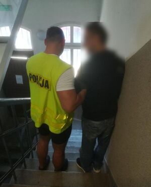 Na zdjęciu widać policjanta, który ma założoną odblaskową kamizelkę koloru żółtego z czarnym napisem POLICJA. Funkcjonariusz prowadzi po schodach w jednostce zatrzymanego mężczyznę.