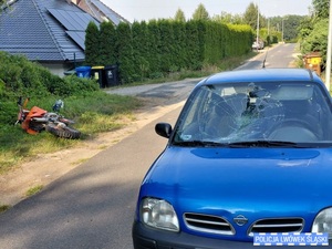 Samochód osobowy z rozbitą przednią szybą zaparkowany przy jezdni. Po lewej stronie na poboczu leży motocykl i kask.