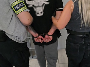 Na zdjęciu widać tyłem trzy osoby. Dwie z nich to policjanci, którzy trzymają za ręce mężczyznę z kajdankami.