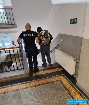 Umundurowany policjant prowadzi mężczyznę, który ma założone kajdanki na ręce trzymane. Obaj mężczyźni są widoczni z tyłu na klatce schodowej.